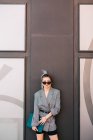 Деловая женщина с модной прической, солнцезащитными очками и костюмом, держащая ноутбук и глядя на камеру на стене — стоковое фото