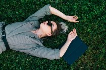 Top view of trendy business woman resting, raising hands and lying on green grass with laptop — стоковое фото