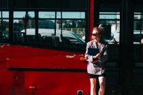 Trendy businesswoman holding tablet, wearing suit and sunglasses, leaning on red bus and looking away — Stock Photo