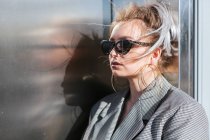 Fashionable attractive woman with trendy hairstyle and sunglasses nearby dark shiny metal wall looking away — Stock Photo