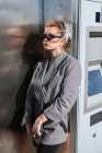 Moda mulher atraente com penteado na moda e óculos escuros nas proximidades parede de metal brilhante escuro olhando para longe — Fotografia de Stock