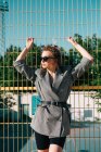 Trendige modische Geschäftsfrau schaut mit Sonnenbrille weg und lehnt an gelbem Gitter bei hellem Tag auf Spielplatz — Stockfoto