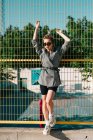 Trendige modische Geschäftsfrau blickt mit Sonnenbrille in die Kamera und lehnt sich bei hellem Tag auf Spielplatz an gelbes Gitter — Stockfoto