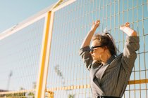 Donna d'affari alla moda guardando lontano in occhiali da sole e appoggiandosi a reticolo giallo in giorno lucente su campo da giuoco — Foto stock