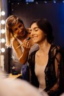 Seitenansicht des Stylisten Make-up auf brünettes Modell sitzt vor beleuchtetem Spiegel in der Umkleidekabine — Stockfoto
