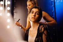 Stylist applying makeup on brunette model sitting in front of illuminated mirror in dressing room — Stock Photo