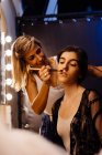 Вид сбоку стилиста, наносящего макияж на модель брюнетки, сидящую перед подсвеченным зеркалом в гримерке — стоковое фото