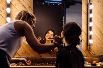 Back view of stylist applying makeup on brunette model sitting in front of illuminated mirror in dressing room — Stock Photo