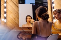 Visão traseira da mulher morena séria aplicando batom vermelho ao fazer maquiagem no vestiário com assistente — Fotografia de Stock