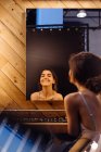 Vista trasera de la joven feliz reflejada en el espejo colgado en la pared de madera mientras se sienta y sonríe en el vestidor - foto de stock