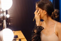 Vue latérale d'une jolie femme aux cheveux foncés en robe transparente noire se maquillant tout en étant assise devant un miroir éclairé — Photo de stock