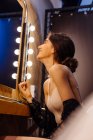 Seitenansicht einer jungen glamourösen Frau, die Lidschatten aufträgt, während sie am beleuchteten Spiegel sitzt und sich in der Garderobe schminkt — Stockfoto