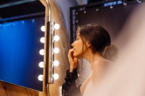 Vista lateral de mujer atractiva con cabello oscuro en vestido transparente negro haciendo maquillaje mientras está sentado frente al espejo iluminado - foto de stock