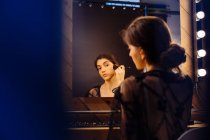 Vista posteriore di donna attraente con i capelli scuri in abito nero trasparente facendo il trucco mentre seduto davanti allo specchio illuminato — Foto stock