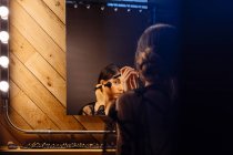 Visão traseira de mulher atraente com cabelo escuro em vestido preto transparente fazendo maquiagem enquanto sentado na frente do espelho iluminado — Fotografia de Stock