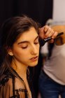 Seitenansicht des Stylisten Make-up auf brünettes Modell sitzt vor beleuchtetem Spiegel in der Umkleidekabine — Stockfoto