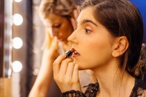 Side view of serious brunette woman in lace black dress applying red lipstick while doing makeup in dressing room — Stock Photo