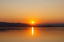 Orange sun going down behind dark hills reflecting in peaceful slightly rippled water creating romantic seascape — Stock Photo