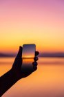Silhueta de um homem tirando uma foto do pôr do sol com seu celular — Fotografia de Stock