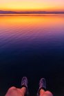 Cultivo piernas de turista irreconocible colgando sobre el agua tranquila del mar durante la puesta del sol brillante - foto de stock