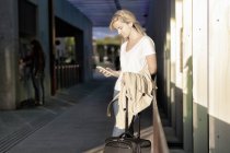 Молода блондинка-туристка в повсякденному одязі використовує смартфон, стоячи в аеропорту з багажем після прибуття в місто — стокове фото