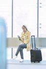 Junge blonde Touristin benutzt Smartphone, während sie mit Gepäck am Flughafen sitzt — Stockfoto