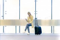 Junge blonde Touristin benutzt Smartphone, während sie mit Gepäck am Flughafen sitzt — Stockfoto