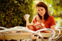 Доросла спокійна концентрована дама читає книгу і п'є чай, сидячи на затишній терасі літнього кафе і насолоджуючись сонячним днем — стокове фото