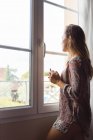 Ragazza bionda pensosa in camicia con una tazza di caffè che guarda fuori le finestre al mattino — Foto stock
