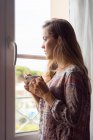 Ragazza bionda pensosa in camicia con una tazza di caffè che guarda fuori le finestre al mattino — Foto stock