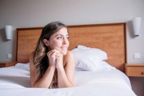 Una chica rubia sonriente en una cama con sábanas blancas iluminadas por la luz de la ventana por la mañana mirando hacia otro lado. - foto de stock