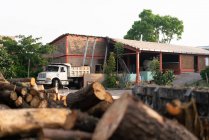 Современный кирпичный дом рядом со старым грузовиком и грудой бревен, окруженных деревьями при дневном свете — стоковое фото