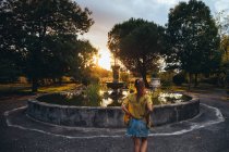 Vista trasera de una mujer bronceada relajándose cerca de una fuente envejecida y mirando el atardecer en el parque de verano - foto de stock