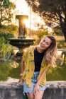 Charmant lächelnde Frau in Freizeitkleidung lehnt am Brunnen bei Sonnenuntergang und blickt in die Kamera im Gegenlicht — Stockfoto