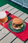 Delicioso cheeseburger caseiro com alface, tomate e molho em prato verde servido com copo de cerveja . — Fotografia de Stock