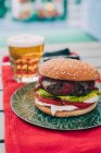 Delizioso hamburger fatto in casa con lattuga, pomodoro e salsa su piatto verde servito con bicchiere di birra . — Foto stock