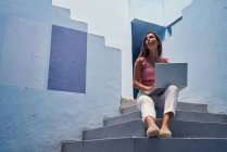 Schöne Frau sitzt auf der blauen Treppe des blauen Gebäudes und benutzt den Computer — Stockfoto