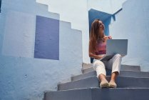 Beautiful woman sitting on blue staircase of blue building and using computer — Stock Photo
