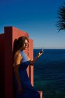 Красивая женщина в синем платье опирается на красную стену здания, держа в руках косметическую бутылку — стоковое фото