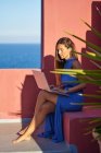 Beautiful barefoot woman in blue dress sitting on step of red building and using laptop — Stock Photo