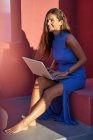 Красива босоніжка в блакитній сукні, що сидить на стадії червоної будівлі і використовує ноутбук — стокове фото