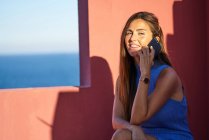 Schöne, glückliche Frau sitzt auf der Stufe des roten Gebäudes und spricht per Smartphone — Stockfoto