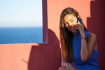 Счастливая женщина сидит на ступеньках красного здания и разговаривает по смартфону — стоковое фото