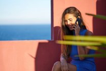 Счастливая женщина сидит на ступеньках красного здания и разговаривает по смартфону — стоковое фото