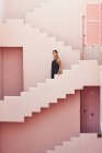 Вид збоку жінки, що йде вниз на сучасну рожеву будівлю, дивлячись далеко — стокове фото