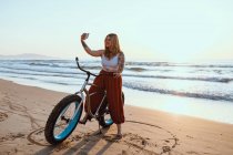 Cheerful chubby woman taking selfie on smartphone while resting with bicycle at sunny shore — Stock Photo