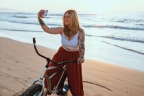 Sorridente contenuto tatuato donna in occhiali scattare foto su smartphone mentre in piedi con la bicicletta in spiaggia tranquilla — Foto stock