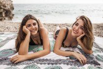 Dos mujeres jóvenes en traje de baño relajándose en la manta en la orilla del mar mirando a la cámara - foto de stock