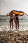 Back view of lesbian couple standing on beach with colorful flag of LGBT movement during summer vacation — Stock Photo