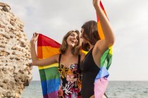 Lesbian couple standing on beach, hugging wrapped in colorful flag of LGBT movement during summer vacation — Stock Photo